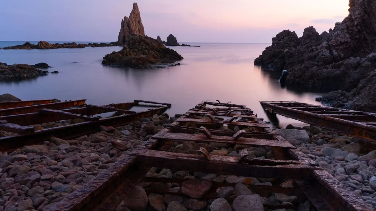 Los pináculos de roca volcánica del Arrecife de las Sirenas saliendo de un mar en calma al anochecer, vistos desde una antigua rampa de barcas oxidada en una playa de cantos rodados de Cabo de Gata