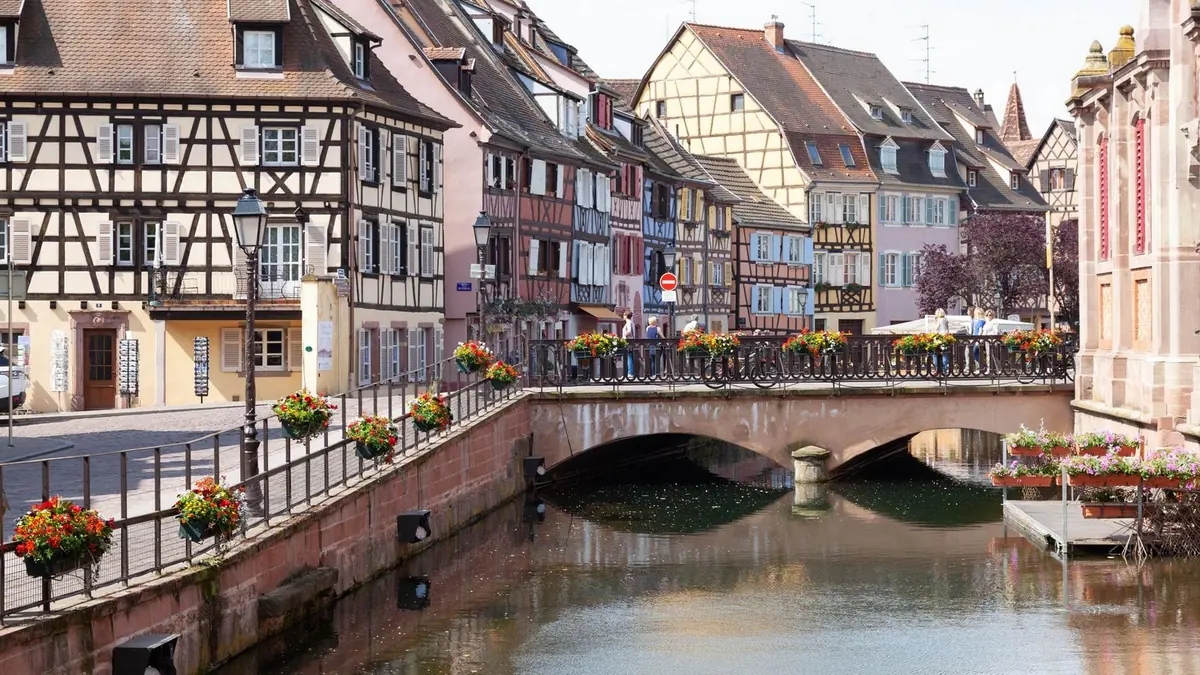 Casas de colores con entramado de madera junto a un canal en Colmar, con un puente de piedra adornado con jardineras de flores rojas y amarillas