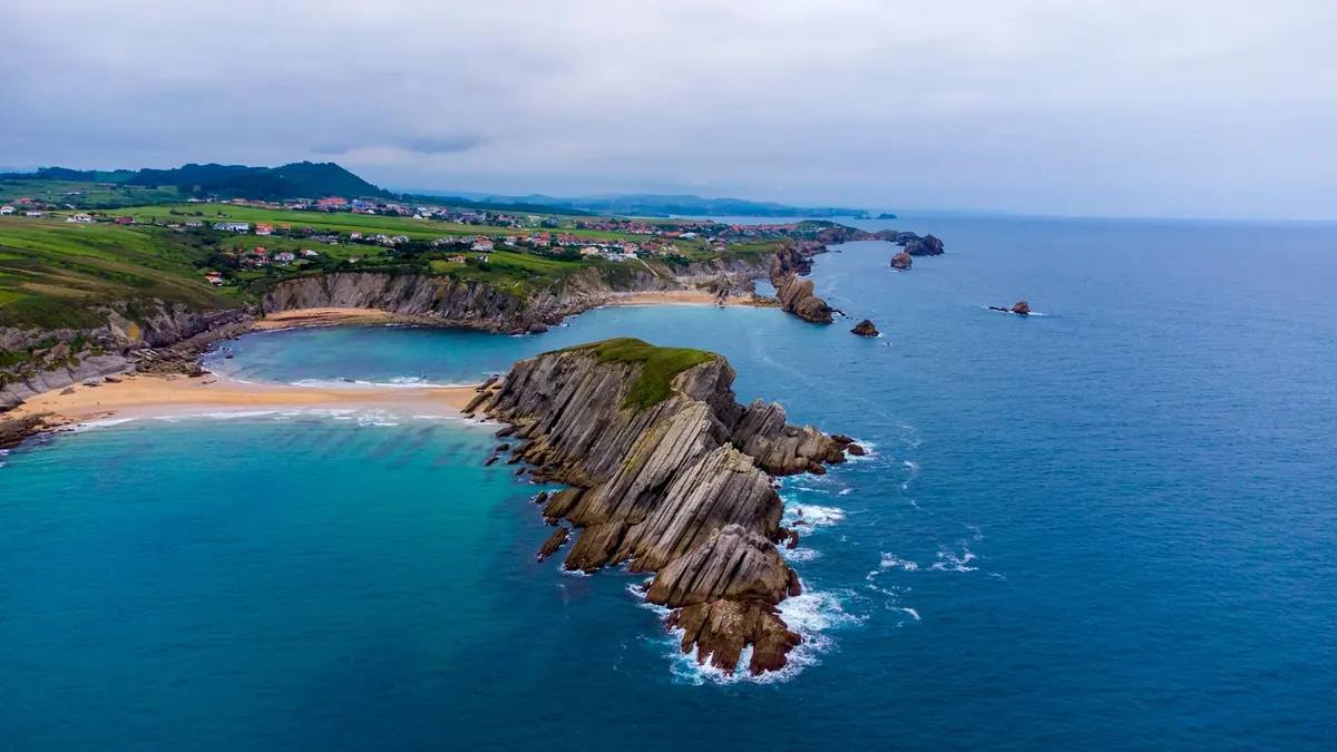 Promontorio di roccia stratificata tra due spiagge di sabbia con acqua turchese e prati verdi sopra le scogliere sulla Costa Quebrada in Cantabria