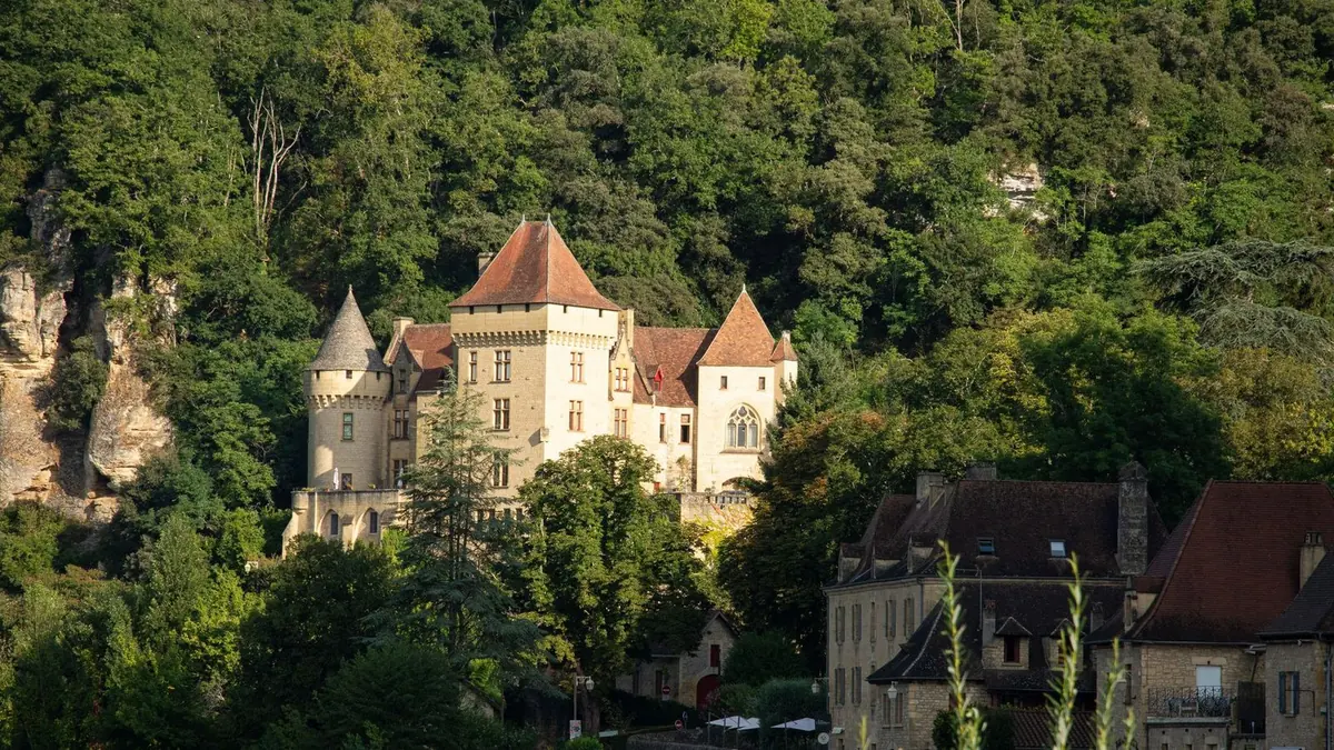 A castle of golden stone with pointed towers and red-tiled roofs below a wooded cliff above the Dordogne at La Roque-Gageac, with old stone houses on the right