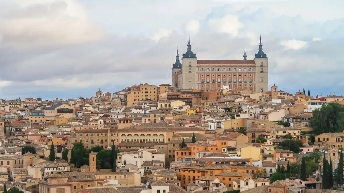 El casco antiguo de Toledo sobre su colina, con casas de piedra y ladrillo y el Alcázar cuadrado de cuatro torres en lo alto, bajo un cielo nublado