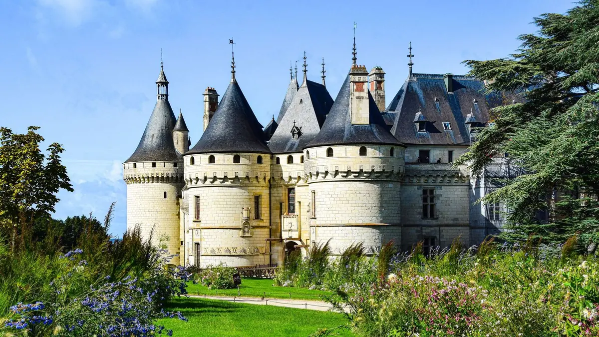 Las torres de piedra blanca y los tejados puntiagudos de pizarra del castillo de Chaumont-sur-Loire tras un césped y parterres con flores azules y rosas