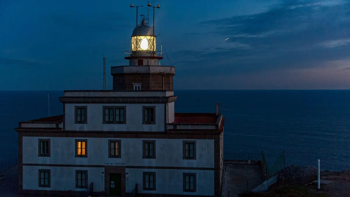 El faro de piedra del cabo Fisterra iluminado al anochecer, con su luz brillando sobre el Atlántico azul oscuro
